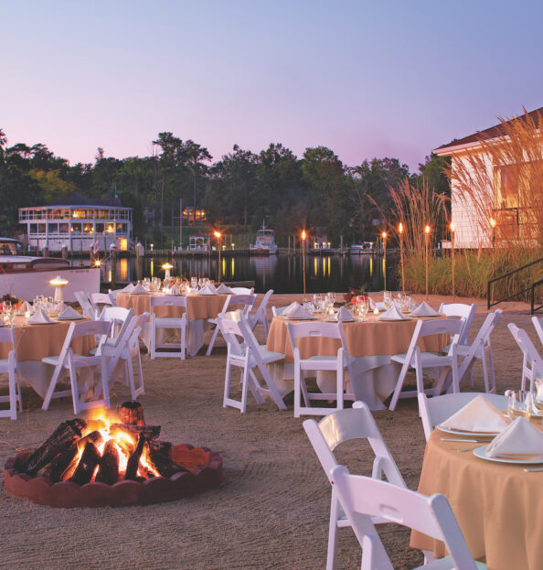 Several tables on the beach set up for fine dining and a fire pit at sunset