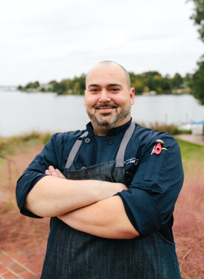 Man in a black chef's coat and apron standing with arms crossed, smiling by the water
