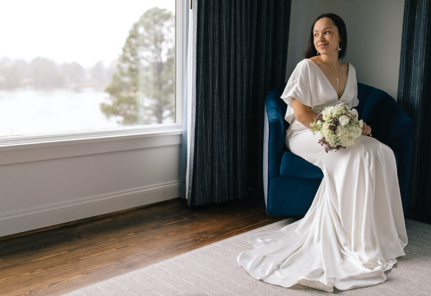 Bride sitting in a royal blue chair by the large window with Chesapeake bay behind her at the Tides Inn